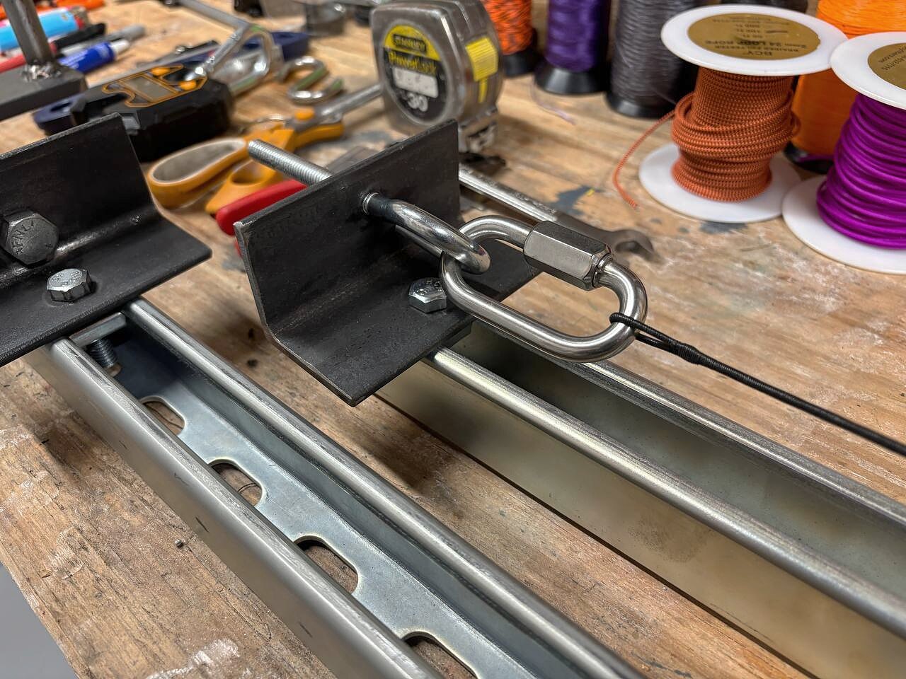 Close-up of the tension end of the string-building jig — a stainless quick-link, threaded rod, and a bracket welded to a slide plate.